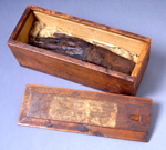 Mummy hand in an open wooden box next to lid with a worn, hand-written label.