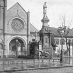 Tall monument topped by sculpture of soldier is flanked by ground-level cannons. In background is a church and several residences