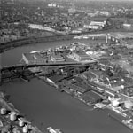 Aerial view of river winding through urban landscape. Bridges, smokestacks, factories, industrial storage tanks, and rowhouse are all visible