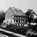 Flat-bed freight cars stand on railroad tracks in foreground. Dilapidated stone building with large open porch stands between tracks and water in background