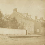 Street-level view of two two-storied buildings with dormer windows. One building is adjacent to fenced tree-filled lot