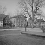 Buildings of diverse architectural styles face fenced-in grass area. Cobblestone and dirt streets run past grassy area