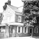 Two-storied stone residence includes dormer windows and first floor windown with open shutters is partially hidden behind large leafy tree. Fenced-in lot sits adjacent to building.