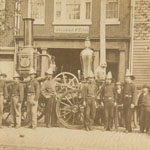 Group of men wearing similar hats and belt buckles pose with wheeled steam engine in front of a brick building. Dog sits on top of steam engine