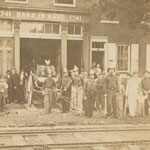 Group of men, some wearing firemen's hats, and boys pose with wheeled fire equipment in front of a brick building. Train tracks run in foreground