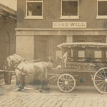 Two horses and a few men pose with a wagon in front of a brick building. Wagon embellished with fire company name and painted images on side