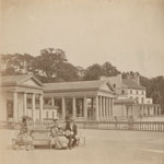 Two women, including one holding an opened parasol, and a man sit on bench near a classical temple-style structure