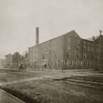 Large multi-storied brick building including tall smoke stack located on street corner. Railroad tracks run down cobblestone street