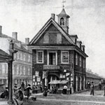 Adjacent to large broadside-covered building is long, one-story structure with many arched entrances. Women with baskets and at tables gather outside main building's front entrance